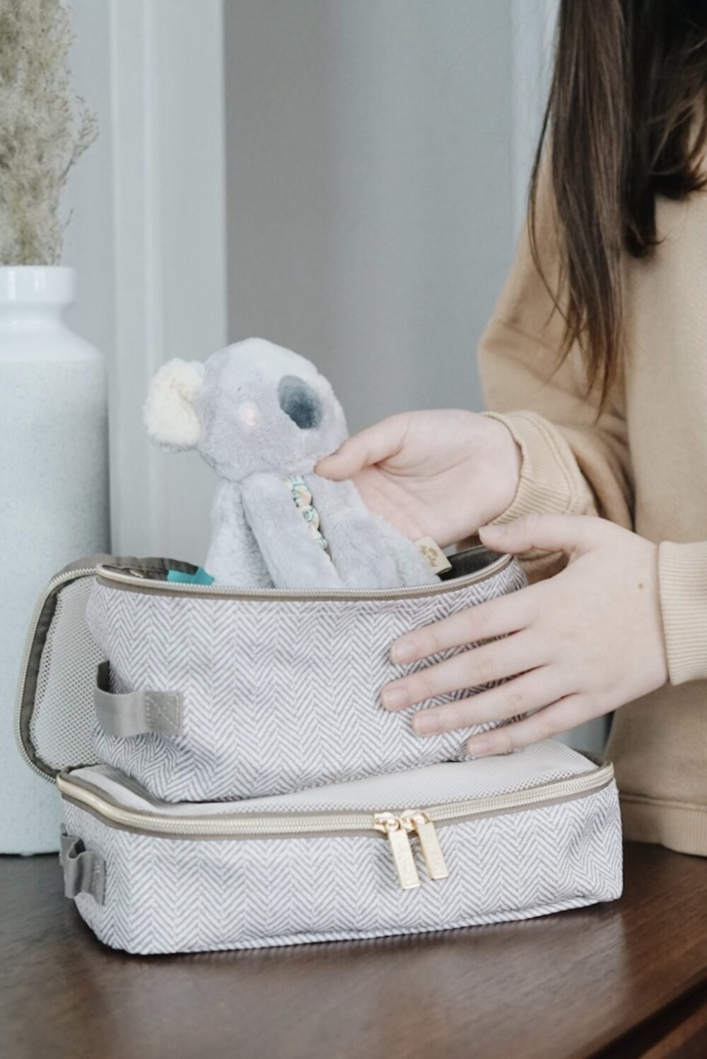 A person is placing a small stuffed koala toy into a gray herringbone-patterned travel case.