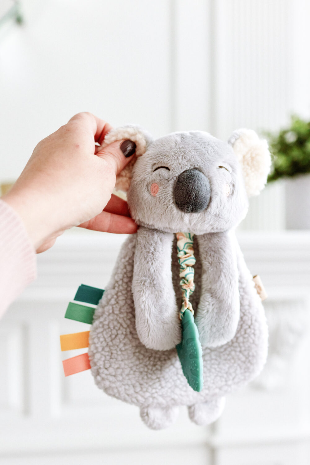 A hand holding a gray, plush koala toy with colorful ribbon tags and a textured blue teething ring, against a light-colored background.