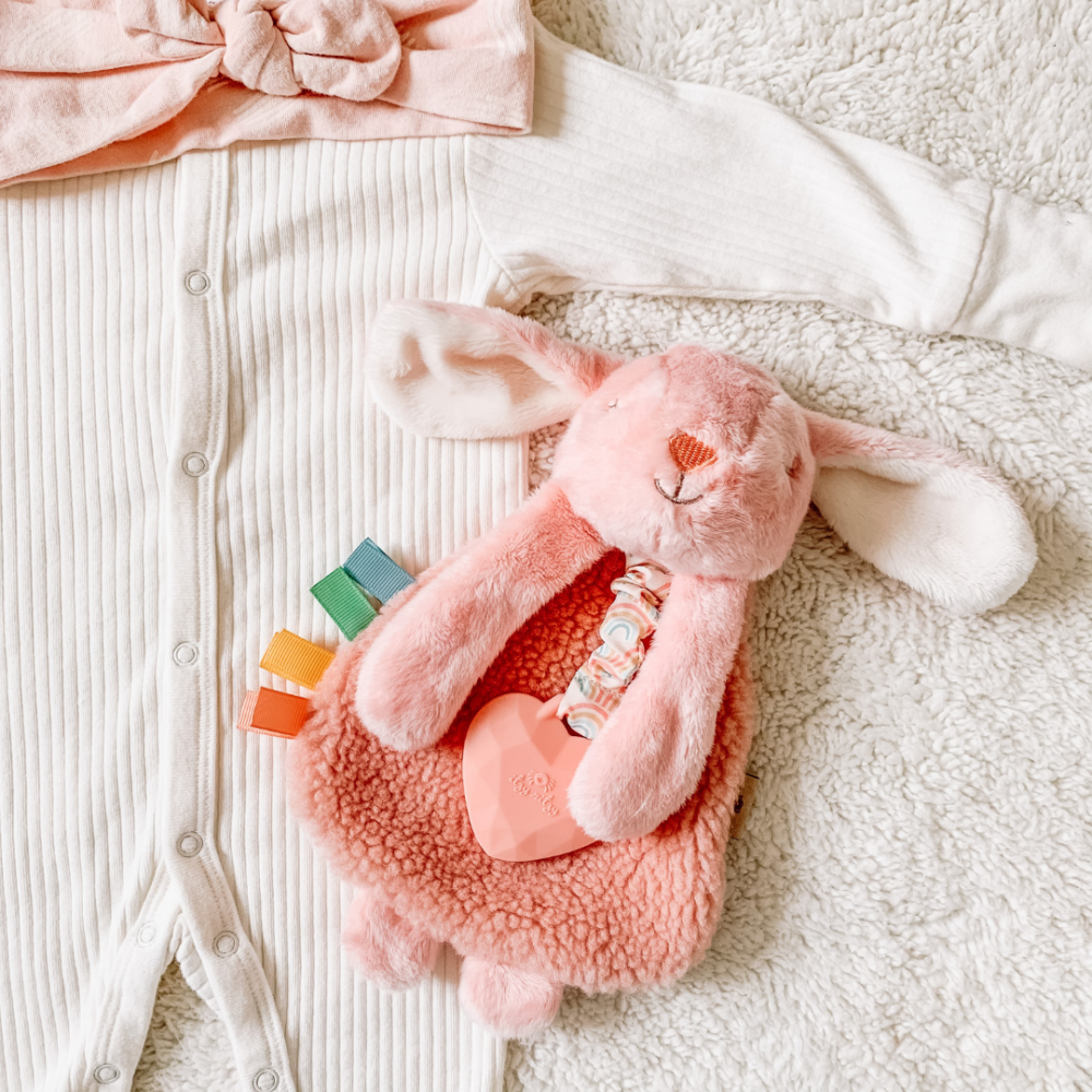 A pink plush rabbit toy and a white ribbed baby onesie with buttons laying on a soft textured surface. A pink headband is partially visible at the top of the image.