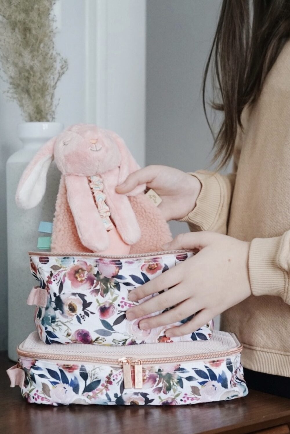 A person is placing a pink stuffed rabbit toy into a floral-patterned bag. Two stacked bags are visible. The background includes a white vase with dried plants.