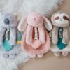 Three plush toys sit side by side on a fluffy white rug. From left to right, they resemble a koala, a pink rabbit, and a sloth. Each toy holds a soft fabric heart and colorful tags.