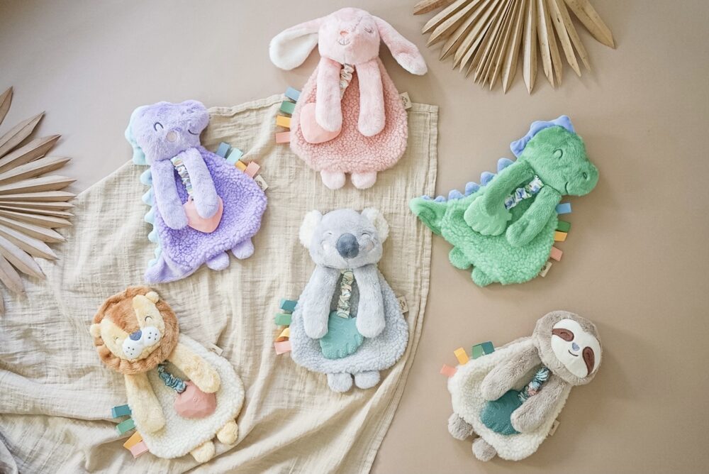 Six plush animal toys, including a rabbit, dinosaur, koala, lion, and sloth, lie on a beige fabric with dried palm leaves in the background. Each toy has a colored trim.