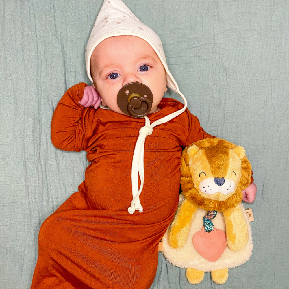 An infant in an orange sleeper and white hat lies on a green blanket, holding a stuffed lion toy. The baby has a pacifier in their mouth.