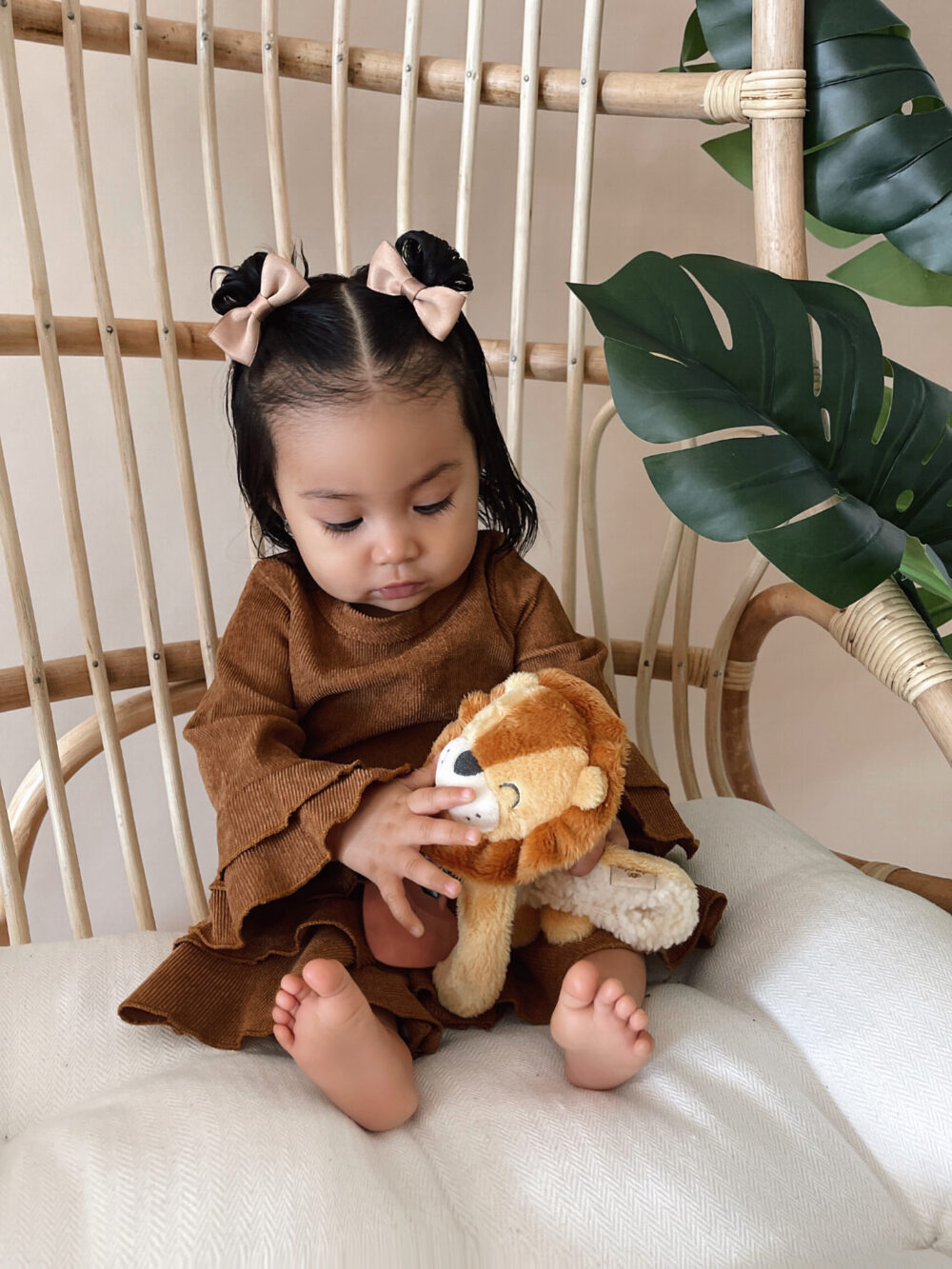 A toddler girl sits on a wicker chair holding a stuffed lion toy, dressed in a brown outfit with hair styled in two buns with bows. Large green leaves decorate the background.