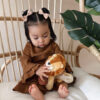 A toddler girl sits on a wicker chair holding a stuffed lion toy, dressed in a brown outfit with hair styled in two buns with bows. Large green leaves decorate the background.