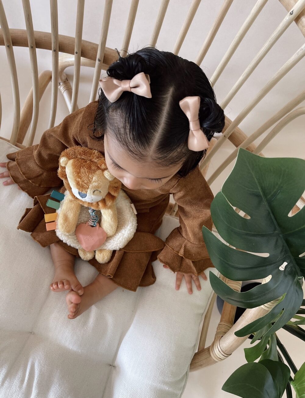 A child with a brown outfit and pink bows in their hair sits on a chair holding a teddy bear, next to a large green leaf.