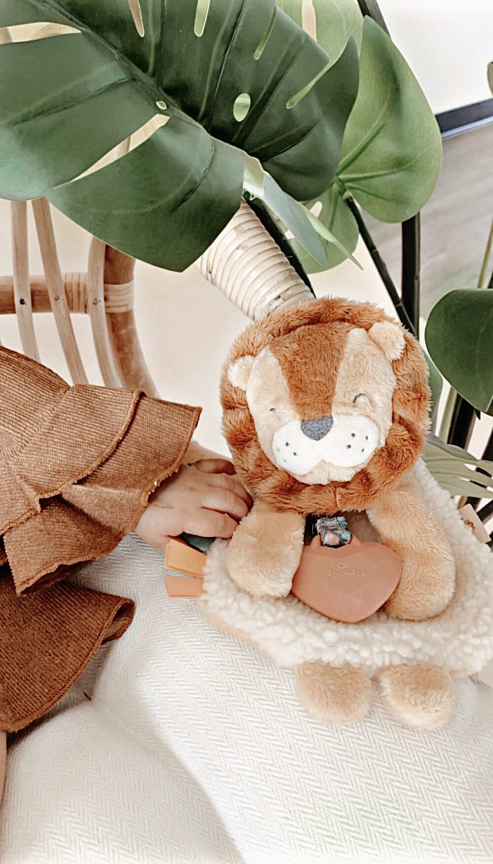 A plush lion toy is placed on a white surface next to a child’s hand and large green leaves. The lion is holding a small object in its front paws.