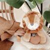 A plush lion toy is placed on a white surface next to a child’s hand and large green leaves. The lion is holding a small object in its front paws.