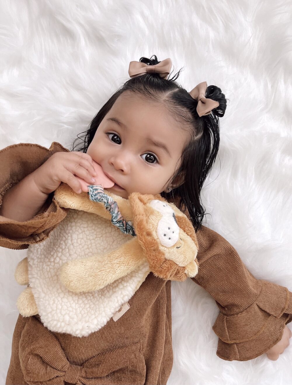 A baby with brown hair and bows lies on a white fluffy surface, wearing a brown outfit and holding a plush lion teether.