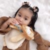 A baby with brown hair and bows lies on a white fluffy surface, wearing a brown outfit and holding a plush lion teether.