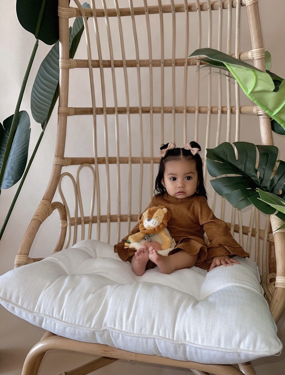 A baby sits on a cushioned wicker chair holding a stuffed lion toy, surrounded by large green leaves.