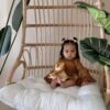 A baby sits on a cushioned wicker chair holding a stuffed lion toy, surrounded by large green leaves.