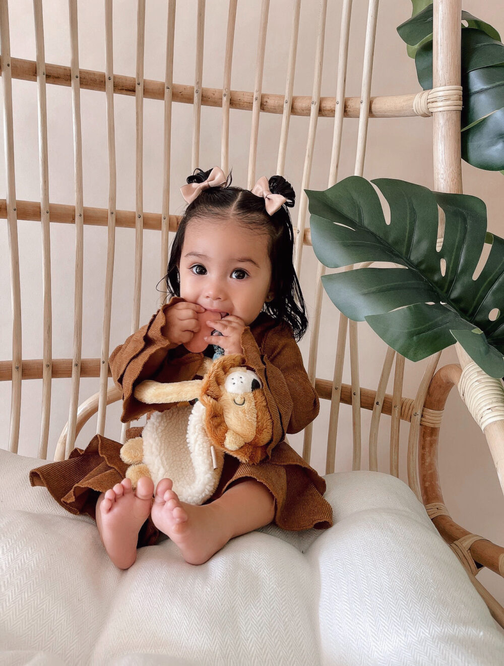 A toddler with two small bows in her hair sits on a wicker chair holding a stuffed animal, surrounded by green plant leaves.