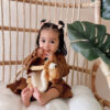 A toddler with two small bows in her hair sits on a wicker chair holding a stuffed animal, surrounded by green plant leaves.