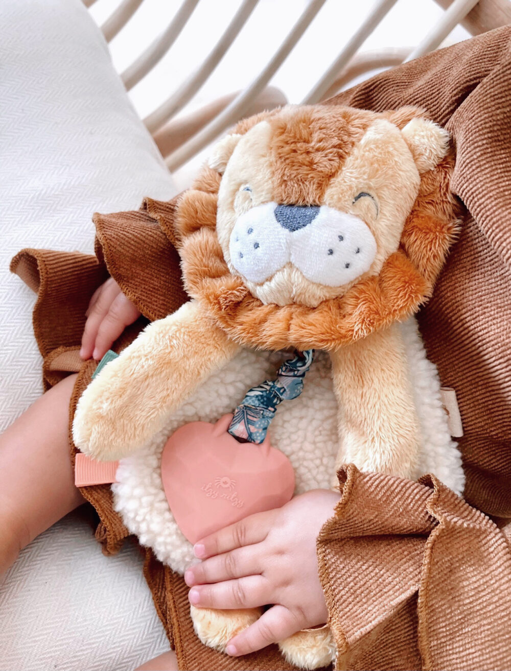 A child holds a plush lion toy with a pink heart-shaped teether and wears a brown outfit with ruffled sleeves.