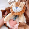 A child holds a plush lion toy with a pink heart-shaped teether and wears a brown outfit with ruffled sleeves.