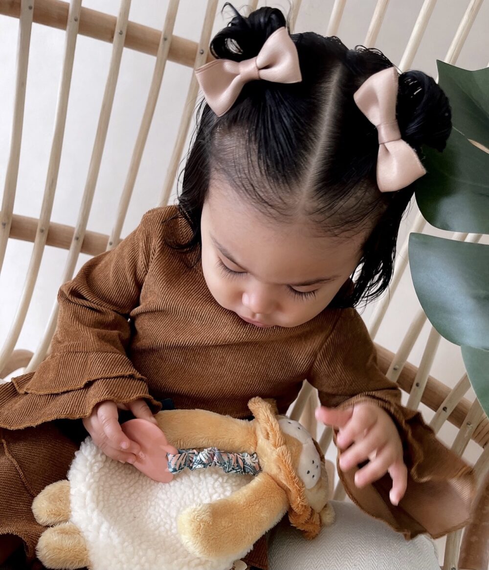 A young child with two bows in their hair, wearing a brown dress, is looking down while holding a stuffed animal. They are sitting on a chair with wooden backrests and next to a green leaf.