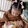 A young child with two bows in their hair, wearing a brown dress, is looking down while holding a stuffed animal. They are sitting on a chair with wooden backrests and next to a green leaf.