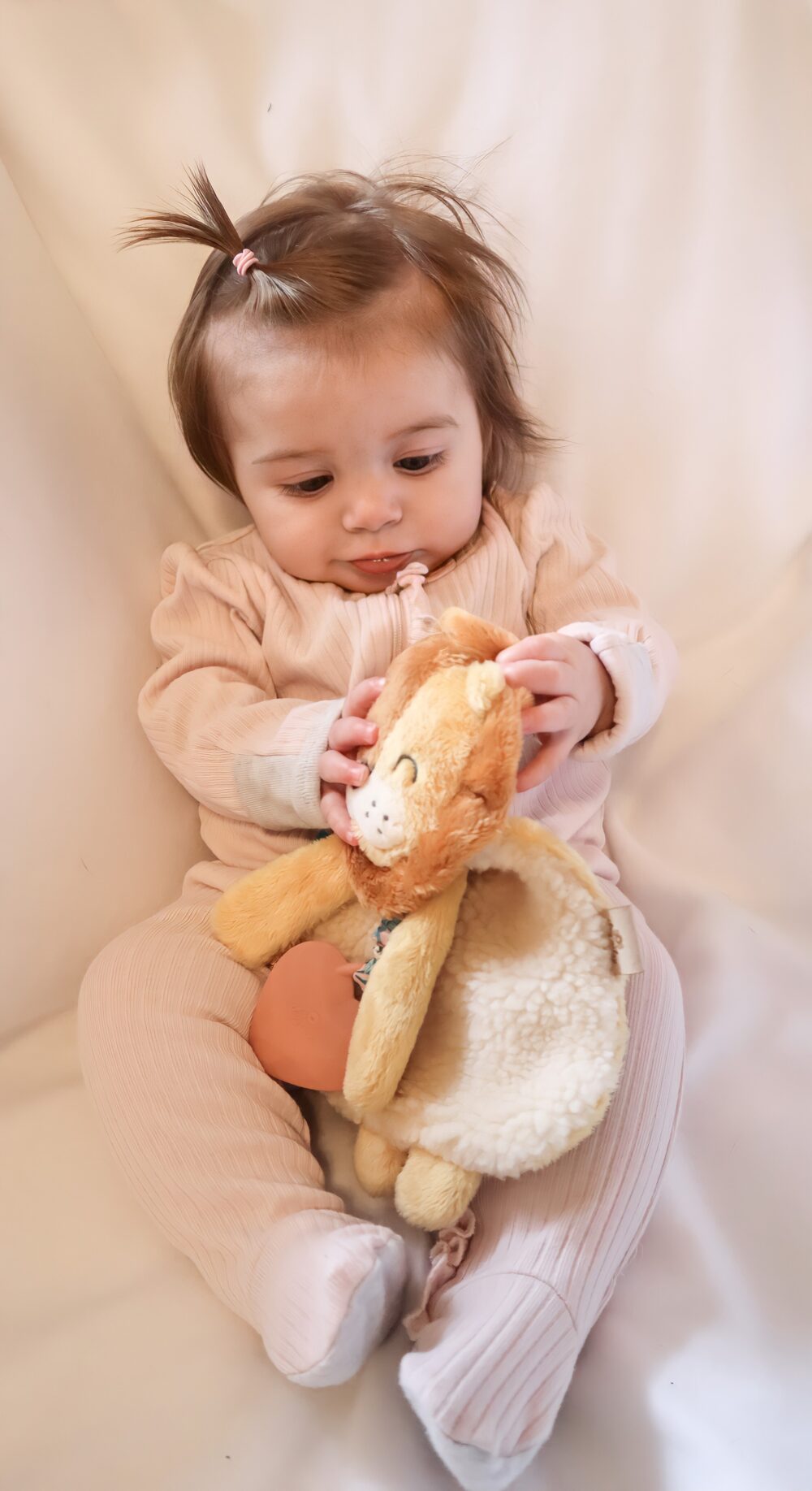 A baby wearing a pink onesie and sitting on a white surface holds and looks at a stuffed lion toy.