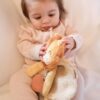 A baby wearing a pink onesie and sitting on a white surface holds and looks at a stuffed lion toy.