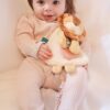 A baby in light pink clothing sits on a white surface, holding a stuffed lion toy.