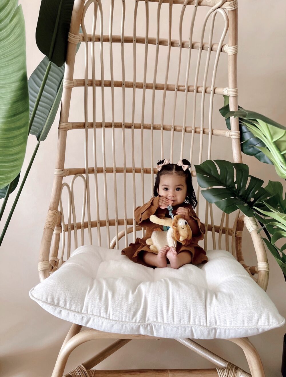 A small child sits on a large wicker chair with a white cushion, holding a plush toy. The chair is surrounded by green plants.