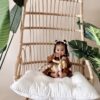 A small child sits on a large wicker chair with a white cushion, holding a plush toy. The chair is surrounded by green plants.