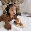 A toddler with bows in her hair and wearing a brown outfit is crawling on a white fluffy rug. She is holding a plush lion toy. Dried brown plants and sheer curtains are in the background.