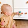 A baby wearing a beige outfit sits on the floor and touches a white dresser drawer with an Itzy Pop & Whirl - Fidget Spinner Highchair, Travel & Bath Toy featuring a smiley face and three colorful balls attached to it.