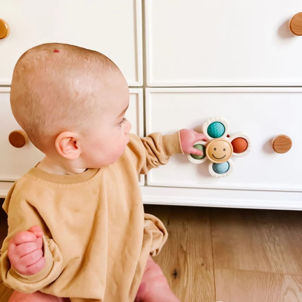 A baby wearing a beige outfit sits on the floor and touches a white dresser drawer with an Itzy Pop & Whirl - Fidget Spinner Highchair, Travel & Bath Toy featuring a smiley face and three colorful balls attached to it.