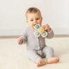 A baby in a grey onesie sits on a beige carpet holding an Itzy Pop & Whirl - Fidget Spinner Highchair, Travel & Bath Toy.