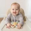 A baby with a bald head in a gray outfit sits in a high chair, smiling and holding an Itzy Pop & Whirl - Fidget Spinner Highchair, Travel & Bath Toy.