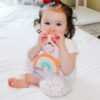 A toddler with curly hair, wearing a rainbow-print shirt and patterned pants, sits on a bed and chews on an Itzy Ritzy - Itzy Pal - Plush Pal Infant Toy with Silicone Teether.