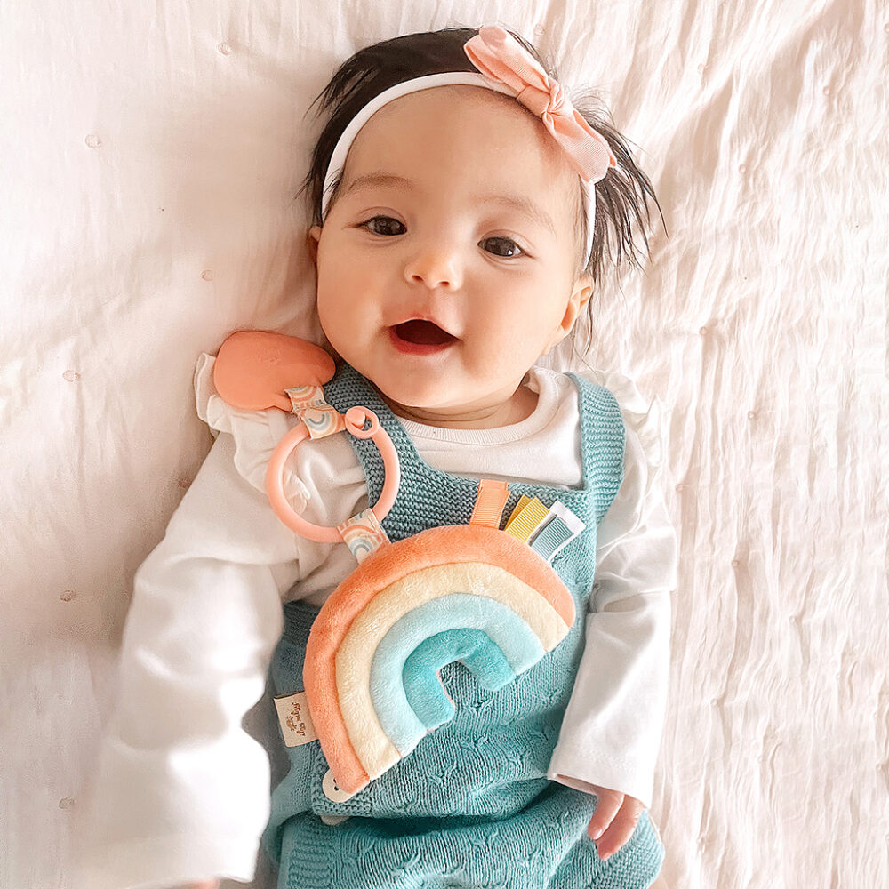 A smiling baby wearing a teal jumper and a pink headband, lying on a white blanket and holding an Itzy Ritzy - Itzy Pal - Plush Pal Infant Toy with Silicone Teether.