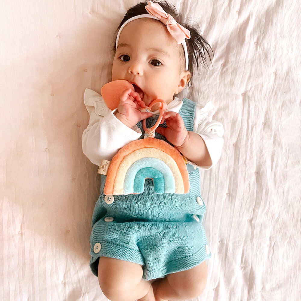 Baby with a headband lying on a pink blanket, chewing on an Itzy Ritzy - Itzy Pal - Plush Pal Infant Toy with Silicone Teether, dressed in a turquoise romper.