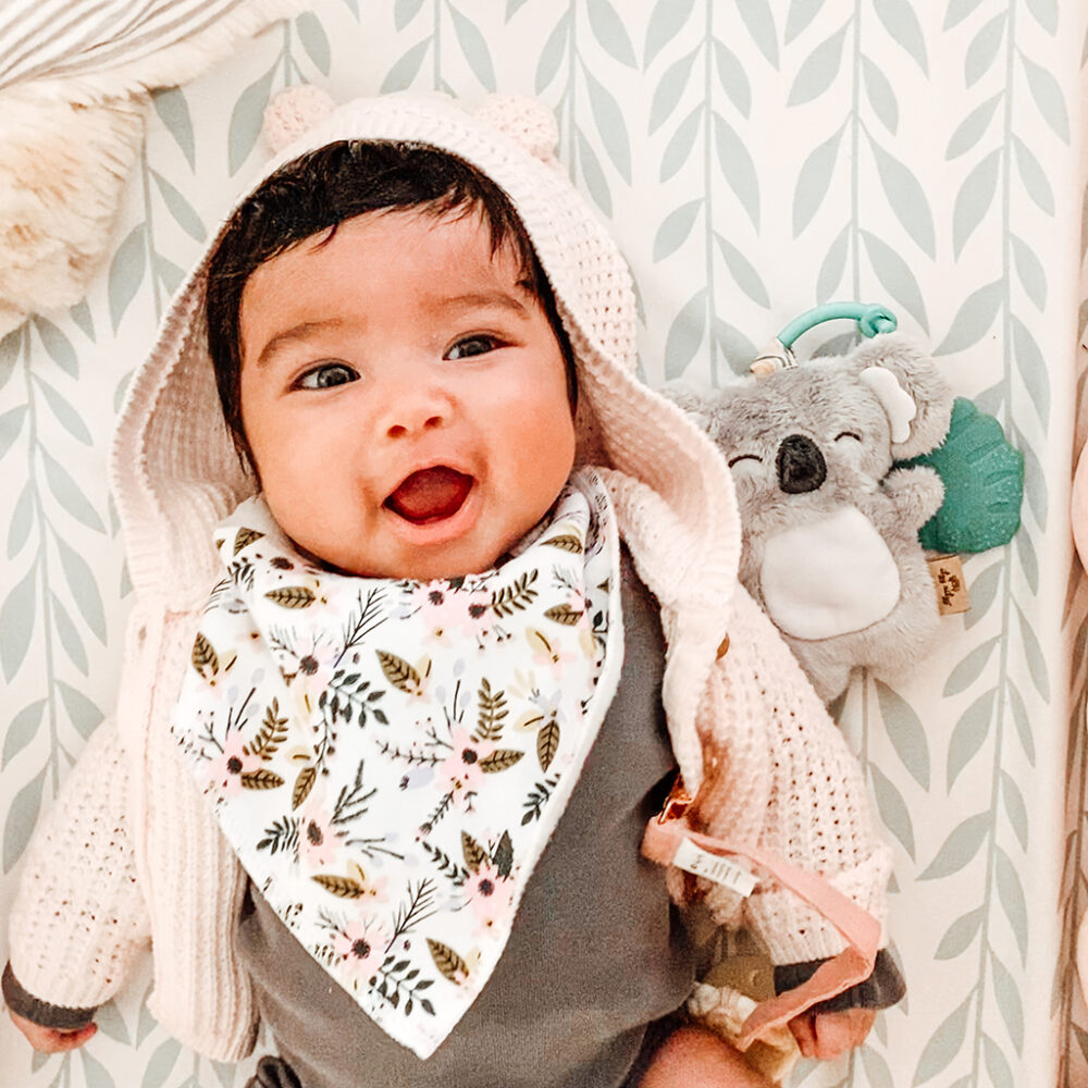 A joyful baby with a floral bib and a bear-eared hood smiles widely, surrounded by soft toys and patterned wallpaper with the Itzy Ritzy - Itzy Pal - Plush Pal Infant Toy with Silicone Teether.