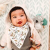 A joyful baby with a floral bib and a bear-eared hood smiles widely, surrounded by soft toys and patterned wallpaper with the Itzy Ritzy - Itzy Pal - Plush Pal Infant Toy with Silicone Teether.