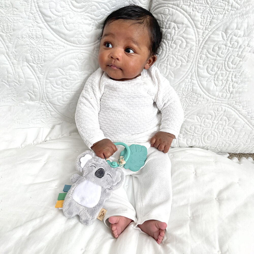 Baby in white outfit sitting on a quilt, holding an Itzy Ritzy - Itzy Pal - Plush Pal Infant Toy with Silicone Teether, looking to the side with a curious expression.