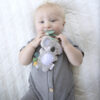 A baby with light hair, wearing a gray onesie, chews on an Itzy Ritzy - Itzy Pal plush koala toy while lying on a white quilted blanket.