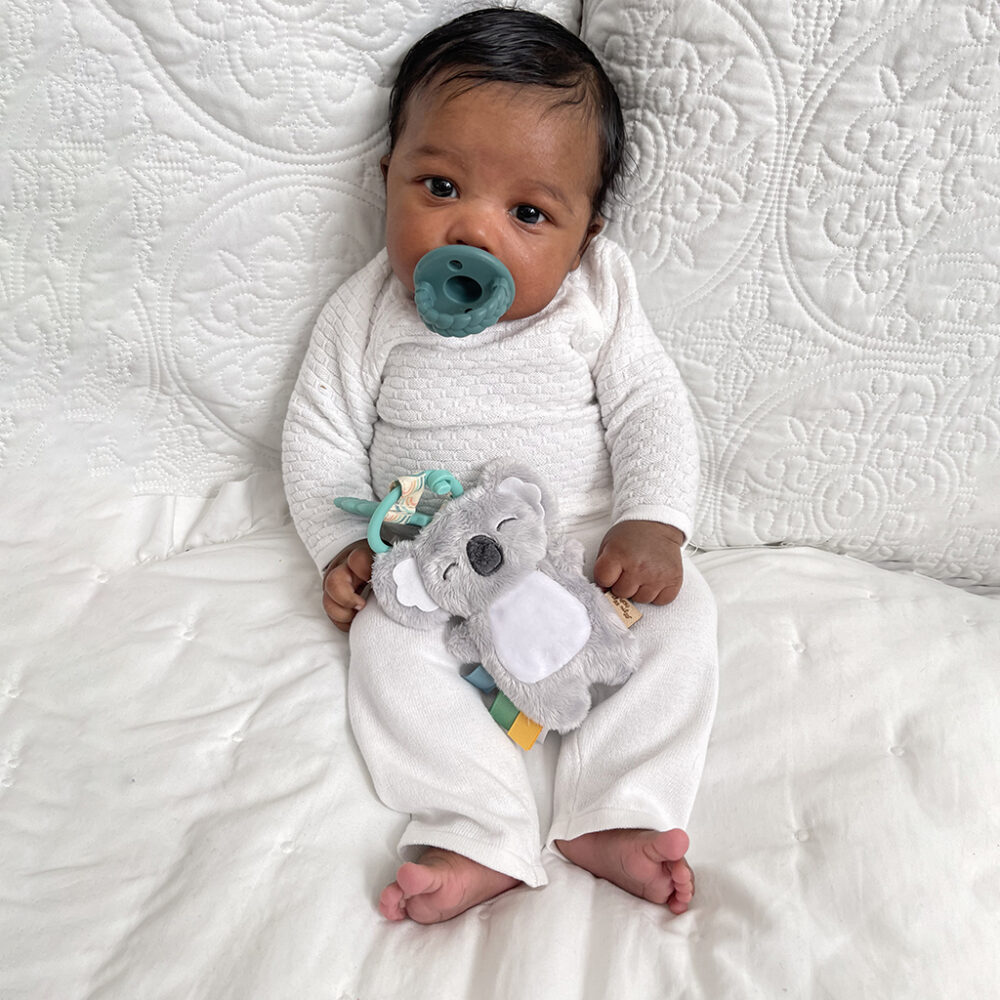 A baby with a pacifier sits on a white quilt, holding an Itzy Ritzy - Itzy Pal - Plush Pal Infant Toy with Silicone Teether.