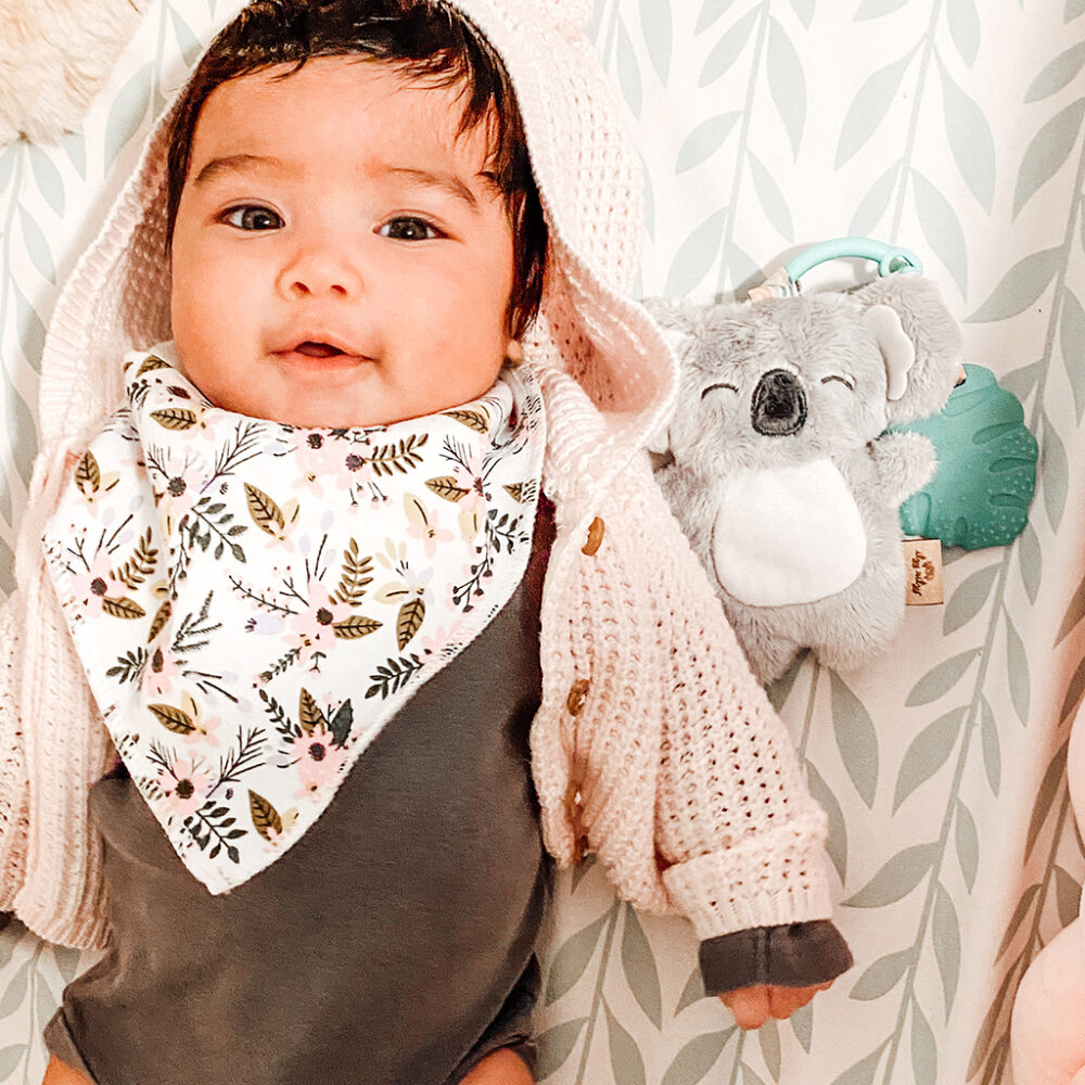 A baby wearing a pink knitted hat and floral bib smiles, lying next to an Itzy Ritzy - Itzy Pal - Plush Pal Infant Toy with Silicone Teether.