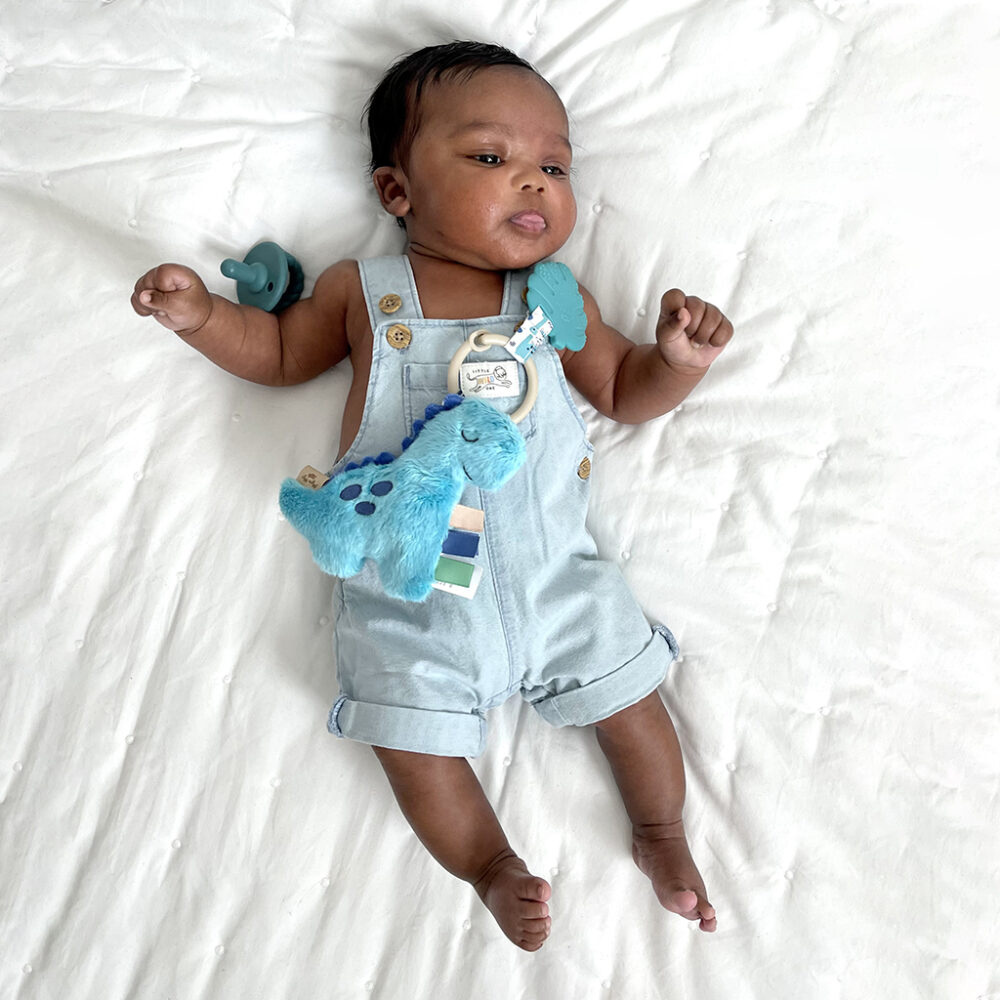 A baby lying on a white sheet, wearing blue overalls and holding an Itzy Ritzy - Itzy Pal - Plush Pal Infant Toy with Silicone Teether.