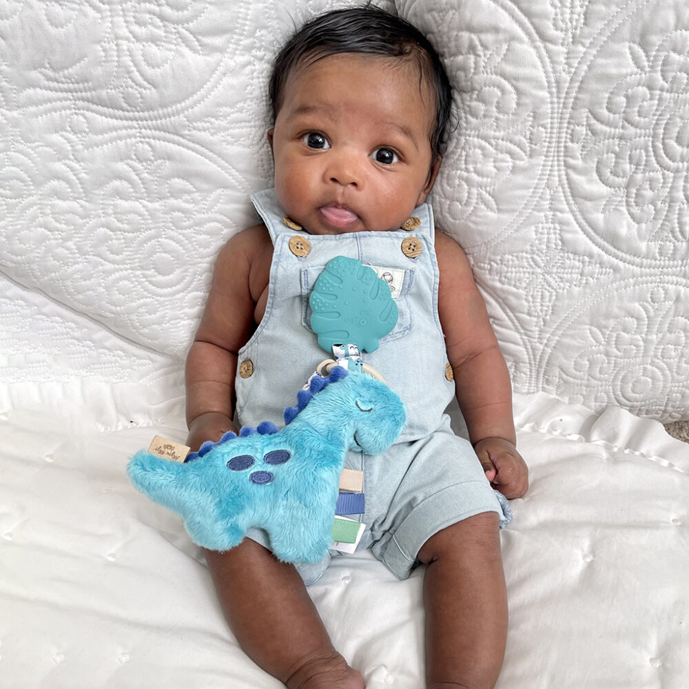 Baby in a denim jumpsuit holding an Itzy Ritzy - Itzy Pal - Plush Pal Infant Toy with Silicone Teether, sitting on a white quilted background.