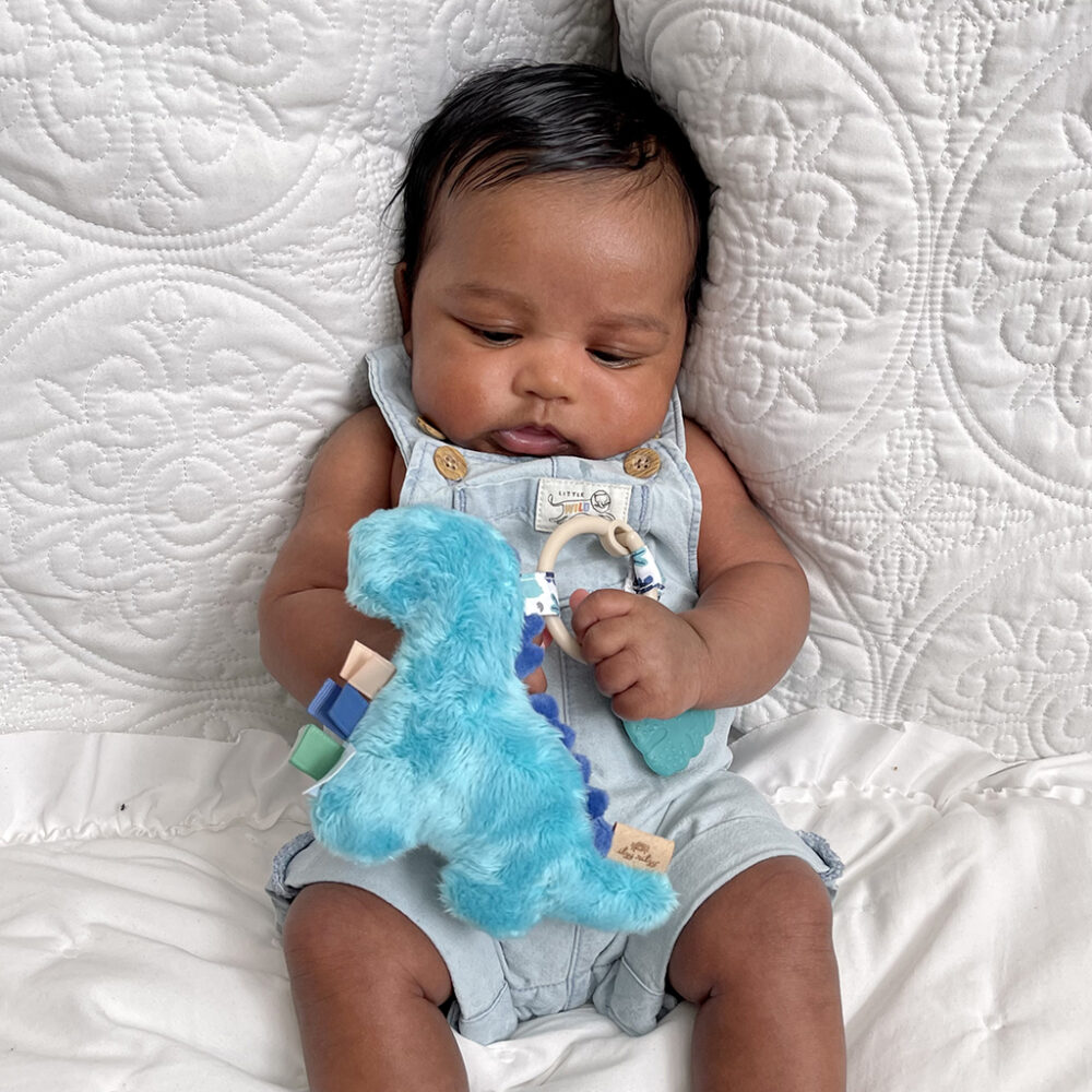 A baby sitting on a textured blanket, holding an Itzy Ritzy - Itzy Pal - Plush Pal Infant Toy with Silicone Teether, dressed in a denim jumper with button details.